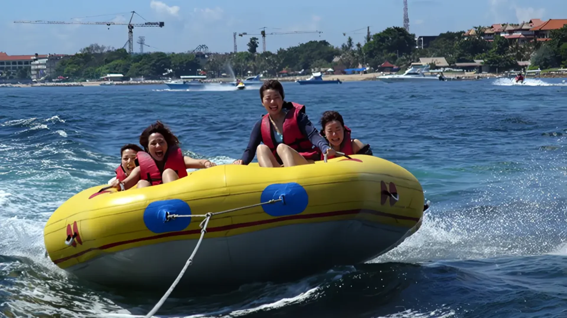 A group of friends smiling and having fun on a rolling donut ride in the choppy waters of Sanur, Bali